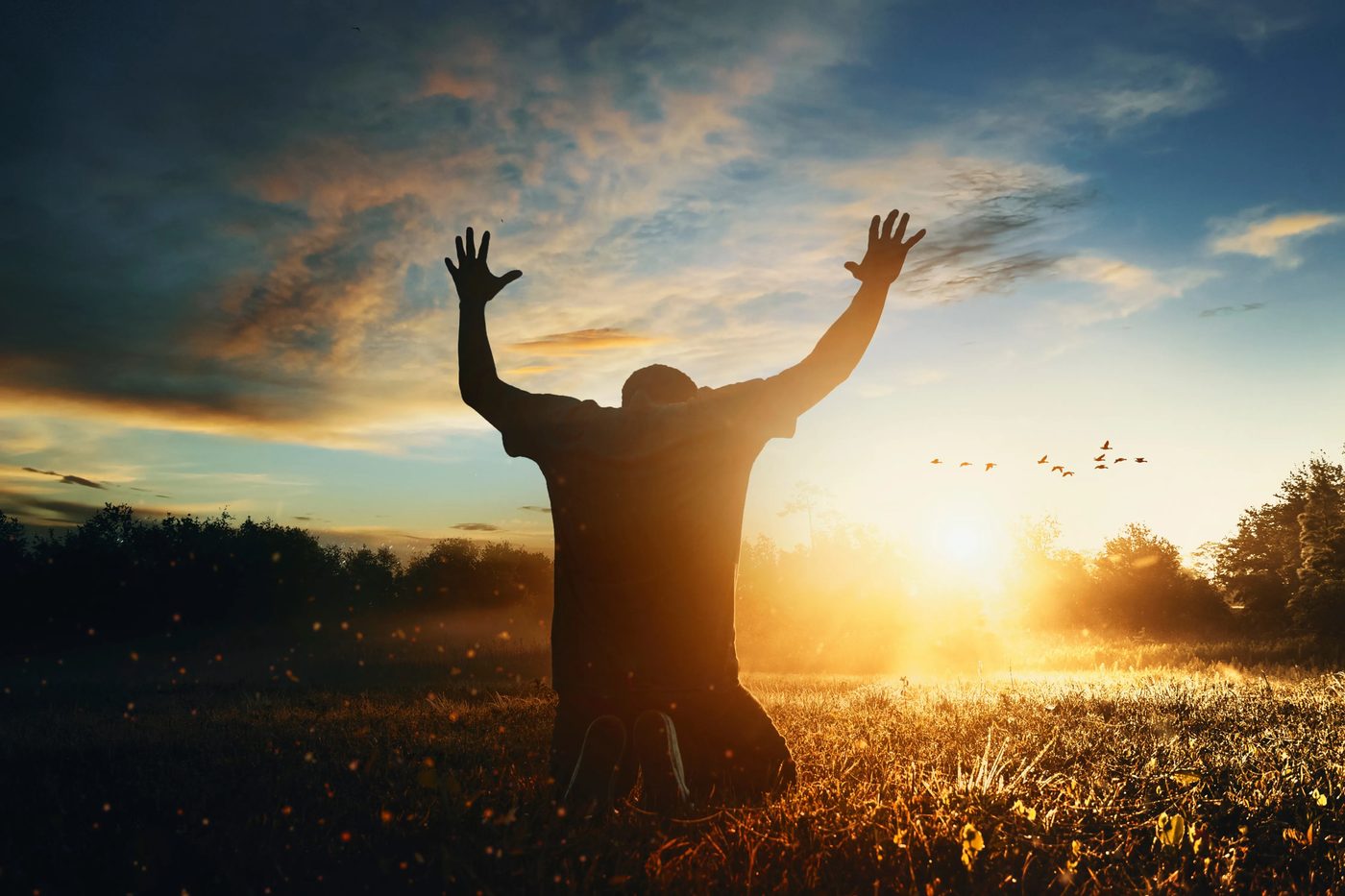 A man kneeling with hands raised in prayer at sunset